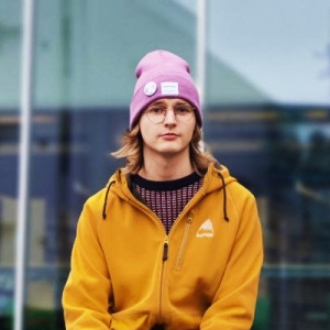 Anders Brandt katsoo kameraan. Häenllä on yllään sinapin värinen takki ja vaaleanpunainen pipo.