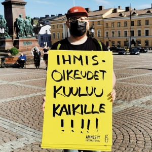 Konsta Hyötylä pitää käsissään kylttiä, jossa lukee "Ihmisoikeudet kuuluu kaikille!!!". Konsta seisoo torilla ja hänellä on musta kasvomaski. 