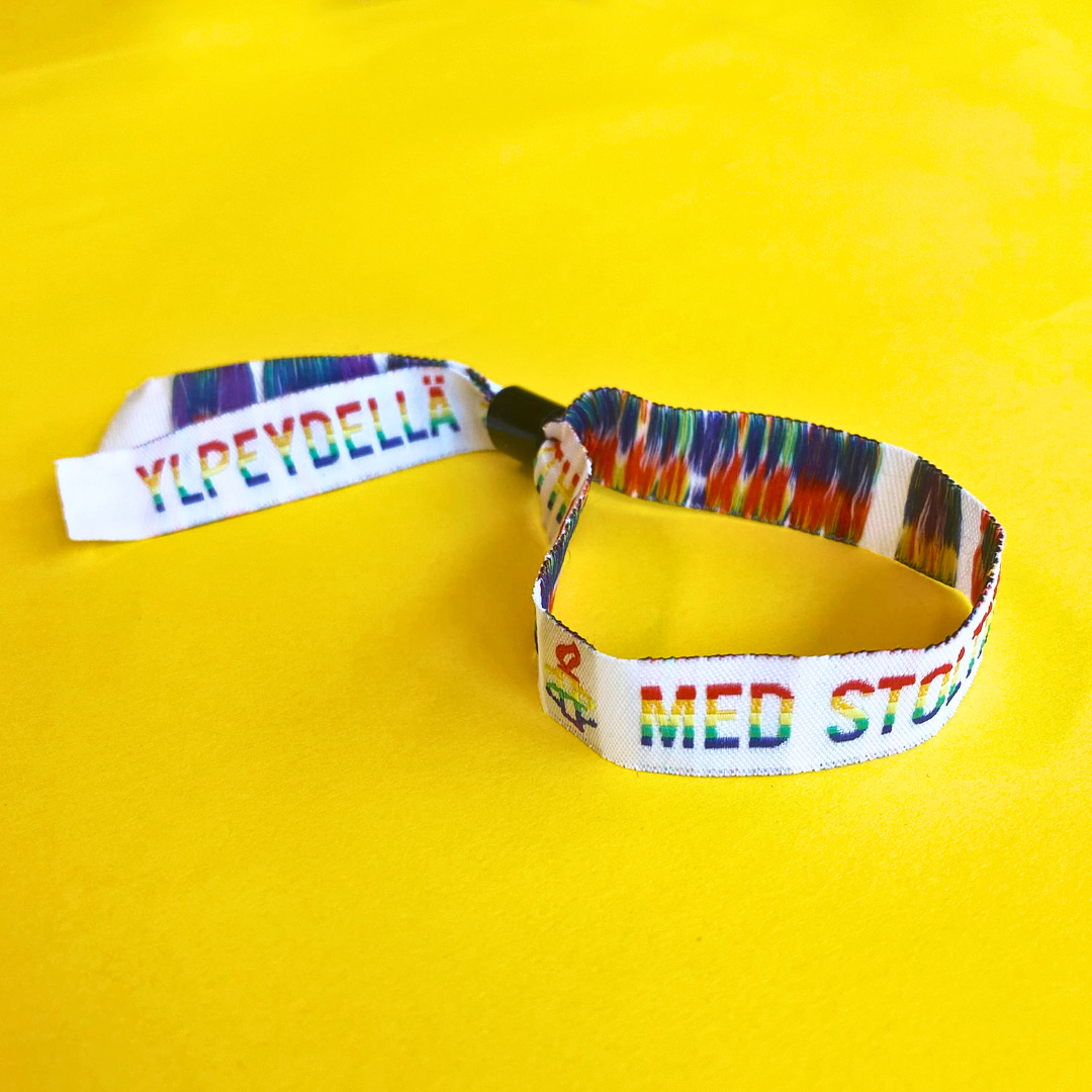 Kudottu kangasranneke, jossa sateenkaaren väreissä fraasi "Ylpeydellä" viidellä eri kielellä.