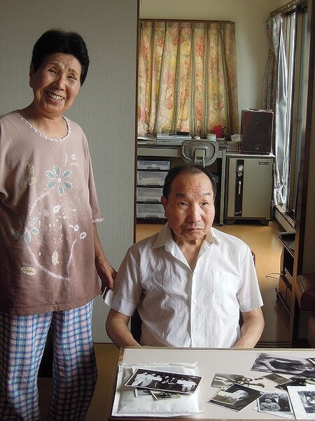 Hideko Hakamada seisoo veljensä Iwao Hakamadan vieressä, joka istuu pöydän ääressä. Pöydällä on vanhoja mustavalkoisia valokuvia. 