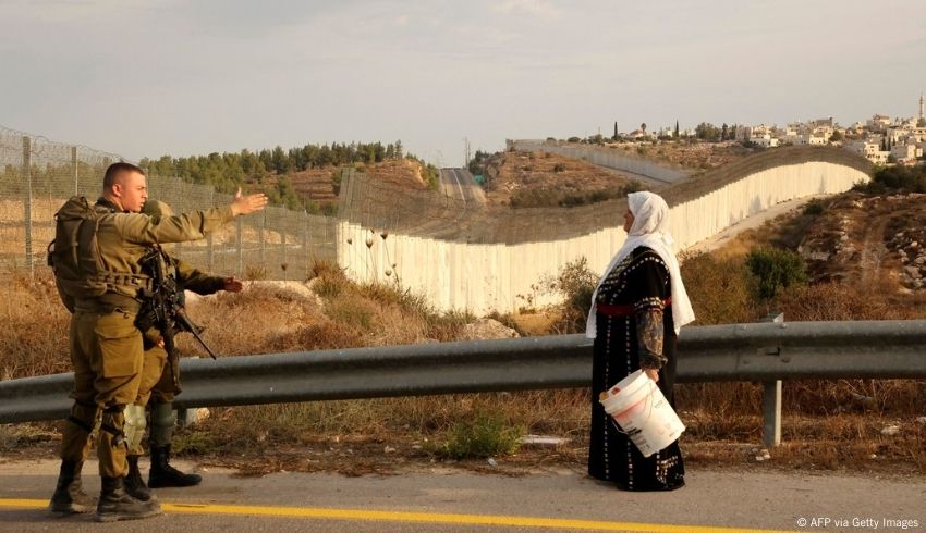 Nainen seisoo tiellä ja pitelee valkoista ämpäriä. Hänen vastassaan on kaksi israelilaista sotilasta.