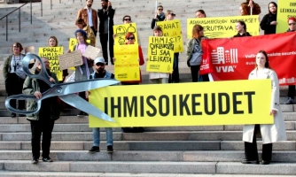 Mielenosoitus eduskuntatalon portailla. Etualalla mielenosoittaja pitää suuria pahvisia saksia, jotka leikkaavat kylttiä, jossa lukee ihmisoikeudet.