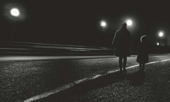 Aikuinen ja lapsi kävelevät tien laitaa pitkin. Kuva on mustavalkoinen.